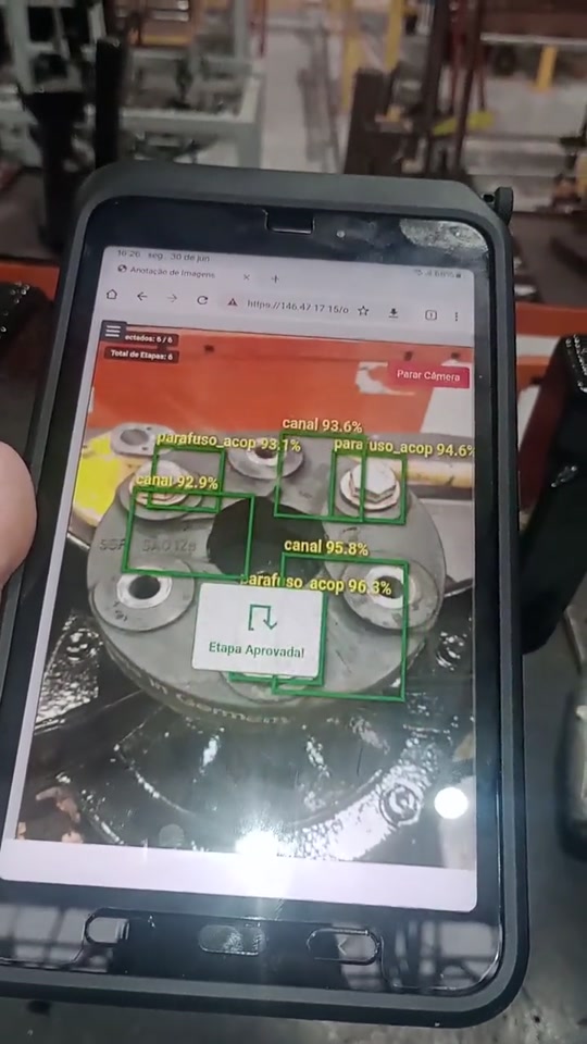 Tablet apontado para um acoplamento montado, com o sistema de visão computacional marcando parafusos e canais em verde e exibindo a mensagem de etapa aprovada